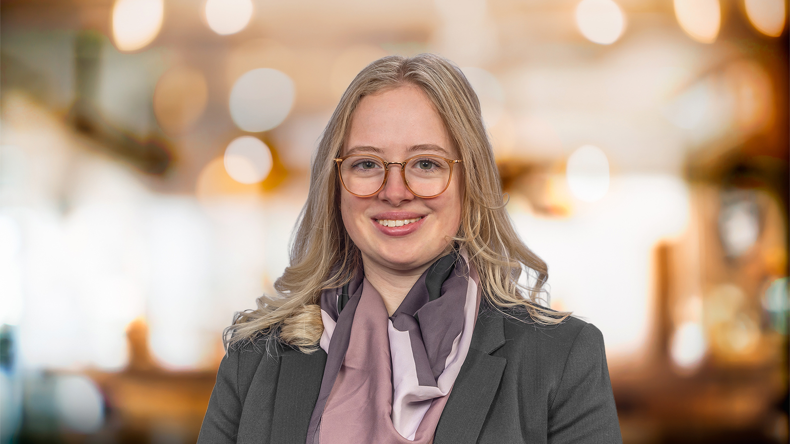 Eine blonde Frau mit Brille und Schal lächelt in die Kamera. Der Hintergrund ist unscharf und warm beleuchtet.